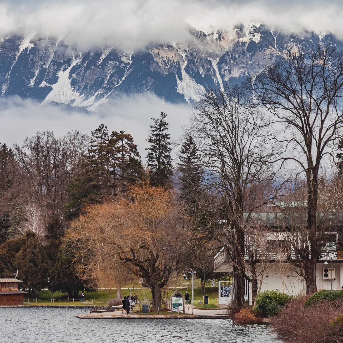 Lakeside path at Lake Bled lined with bare trees and people walking, framed by dramatic clouds clinging to the snowy Julian Alps in the background.
