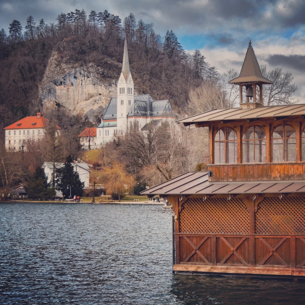 Wooden boathouse on Lake Bled with a view of St. Martin’s Parish Church in the background, nestled at the foot of a rocky hill under a moody sky.