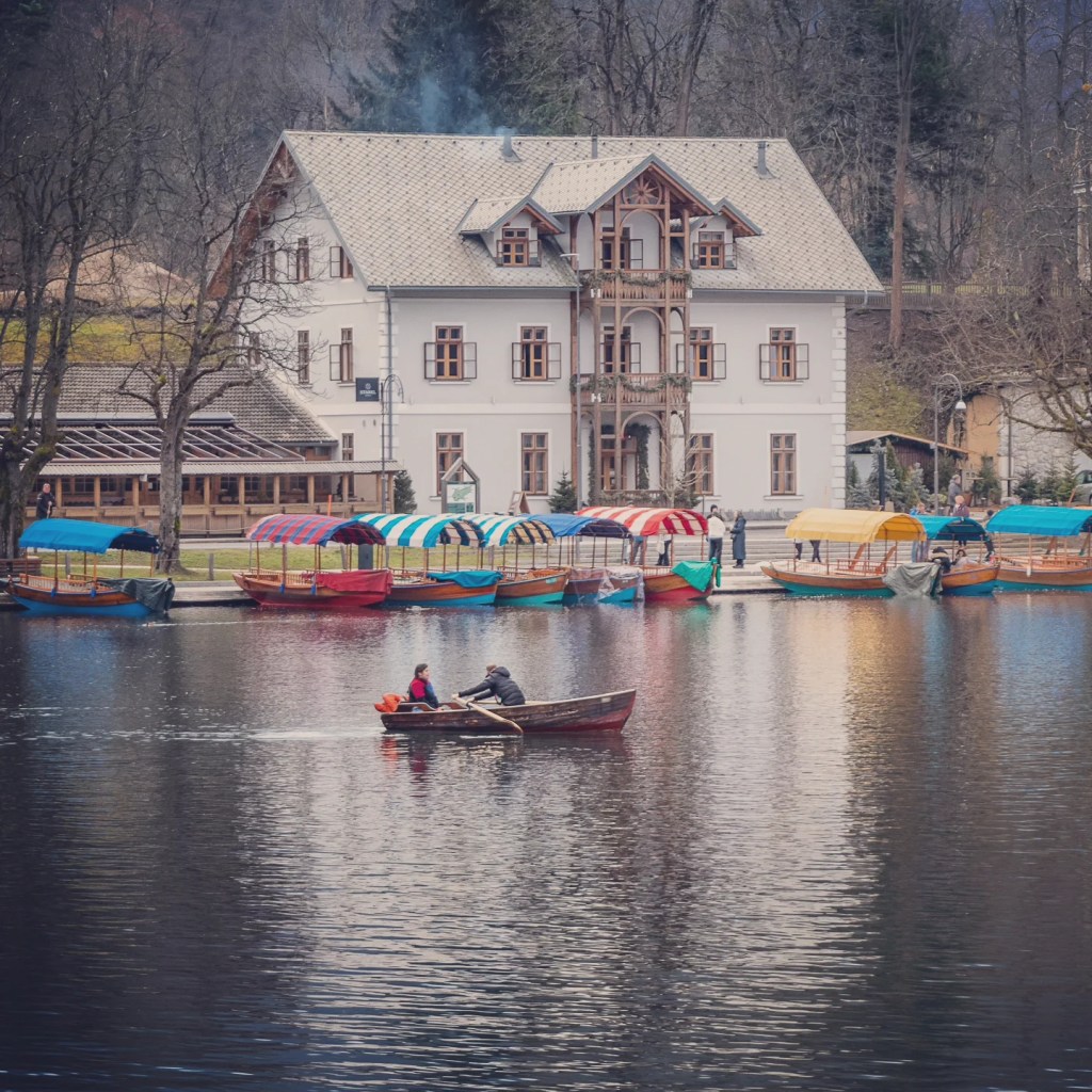 A small rowboat with two people gliding past a row of colorful, covered pletna boats docked along the shore, in front of a charming lakeside villa at Lake Bled.