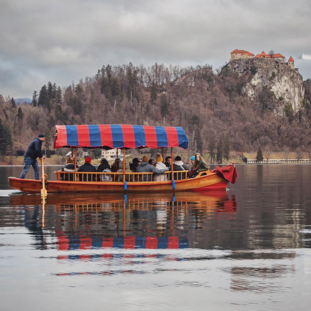 A vibrant wooden pletna boat with a red and blue striped canopy glides across Lake Bled, carrying passengers, with Bled Castle standing on the cliff behind.