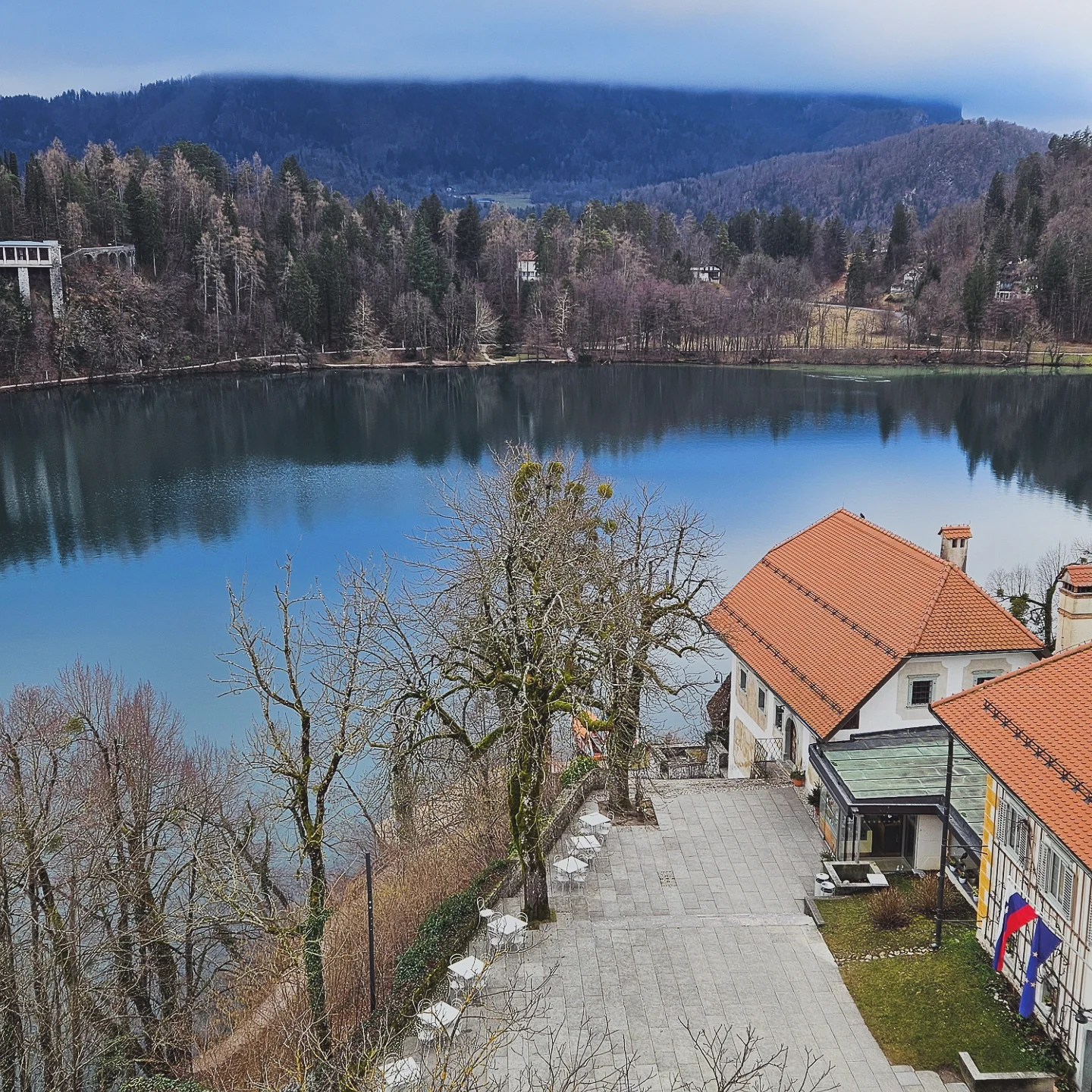 A terrace lined with leafless trees and empty white tables overlooking the glassy surface of Lake Bled, with forested hills rising into misty mountains in the distance.