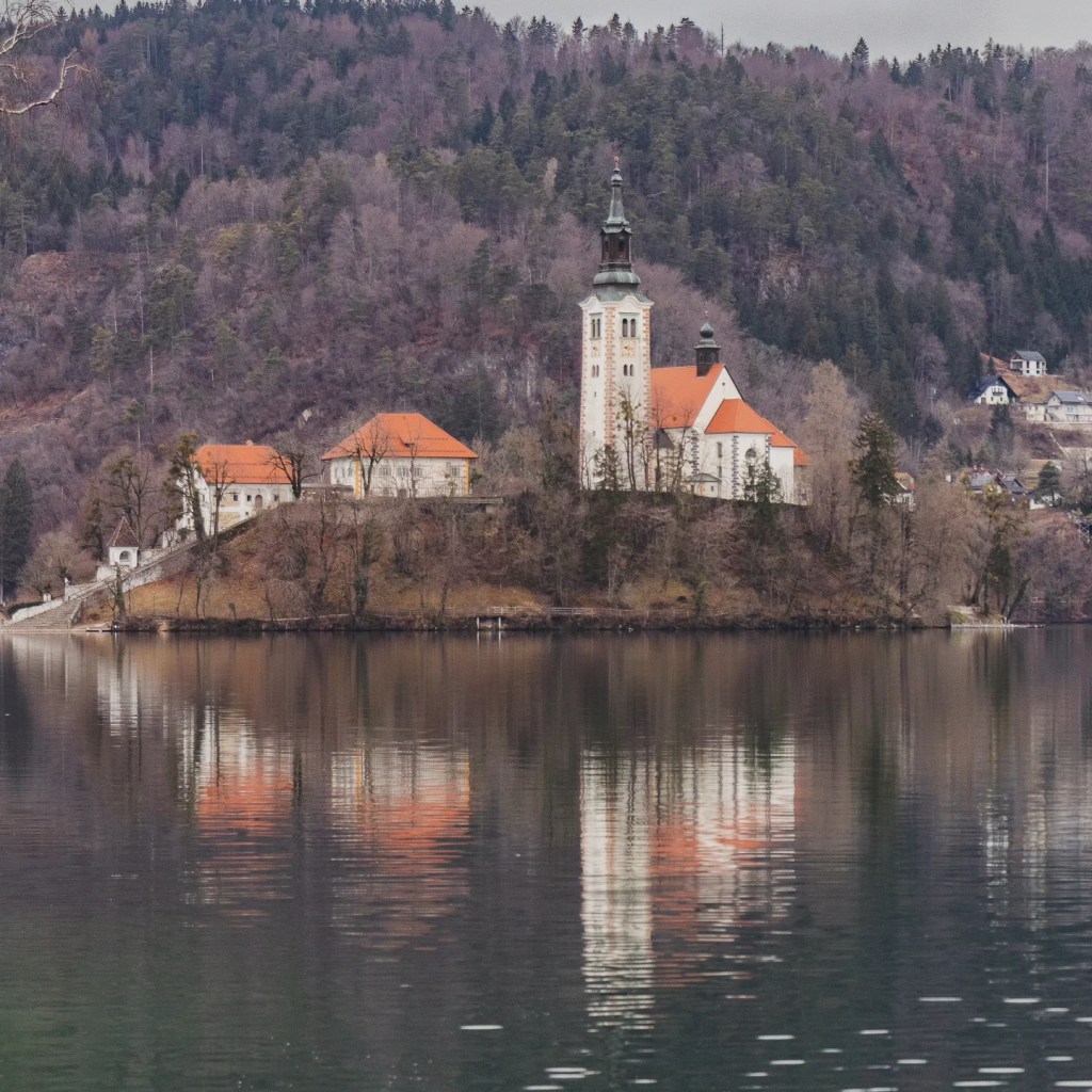 Wide view of the Church of the Mother of God on the Lake on Bled Island reflected in the still waters, surrounded by forested hills with scattered homes.
