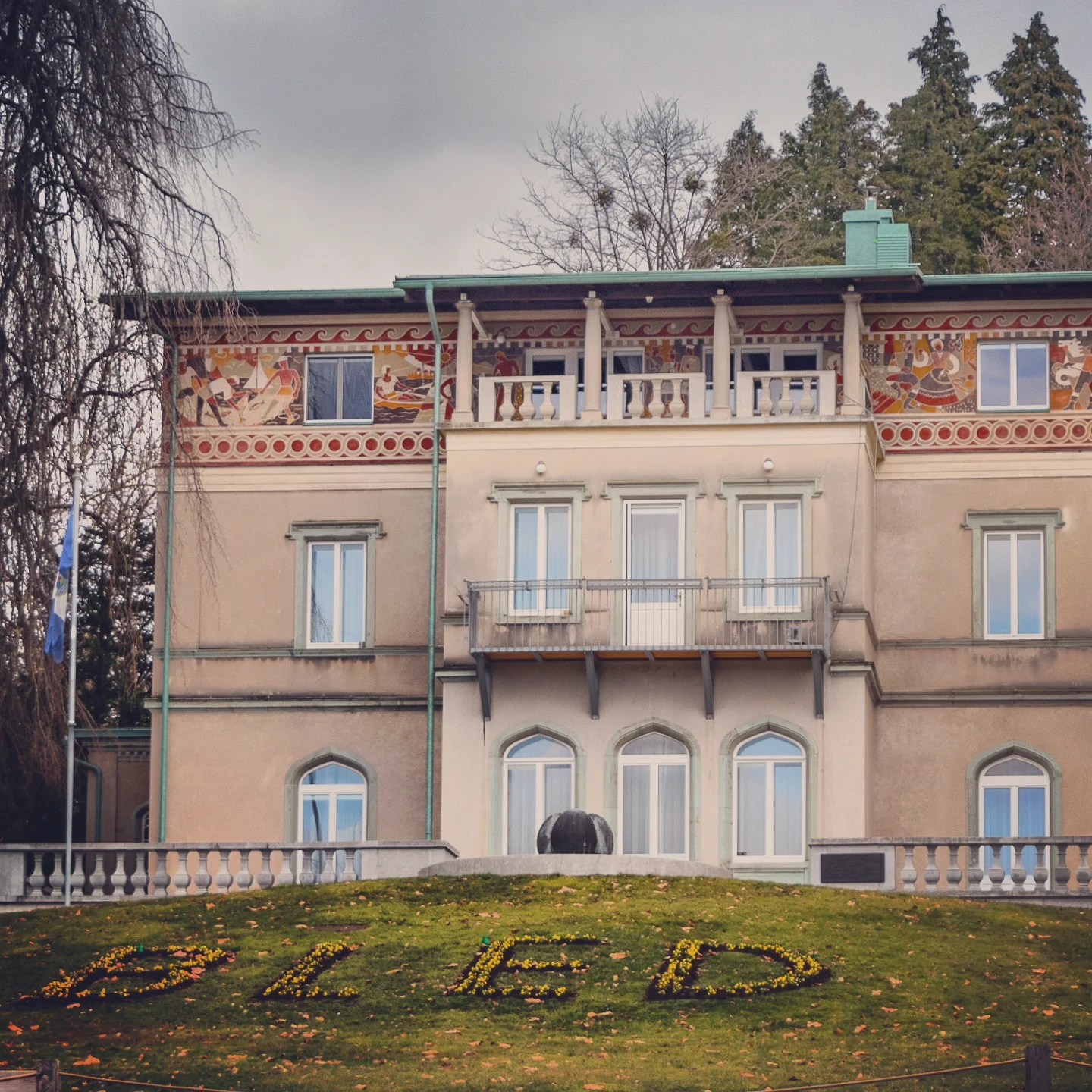Historic building with decorative frescoes above its balcony and the word “Bled” spelled in flowers on the green lawn in front.