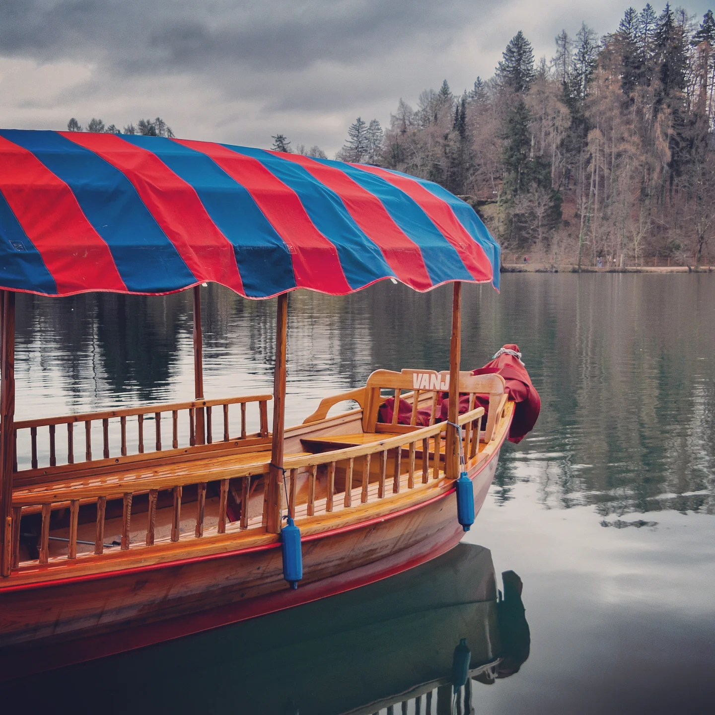 A colorful red and blue striped pletna boat gently docked on the calm waters of Lake Bled, surrounded by forested shoreline and cloudy skies.