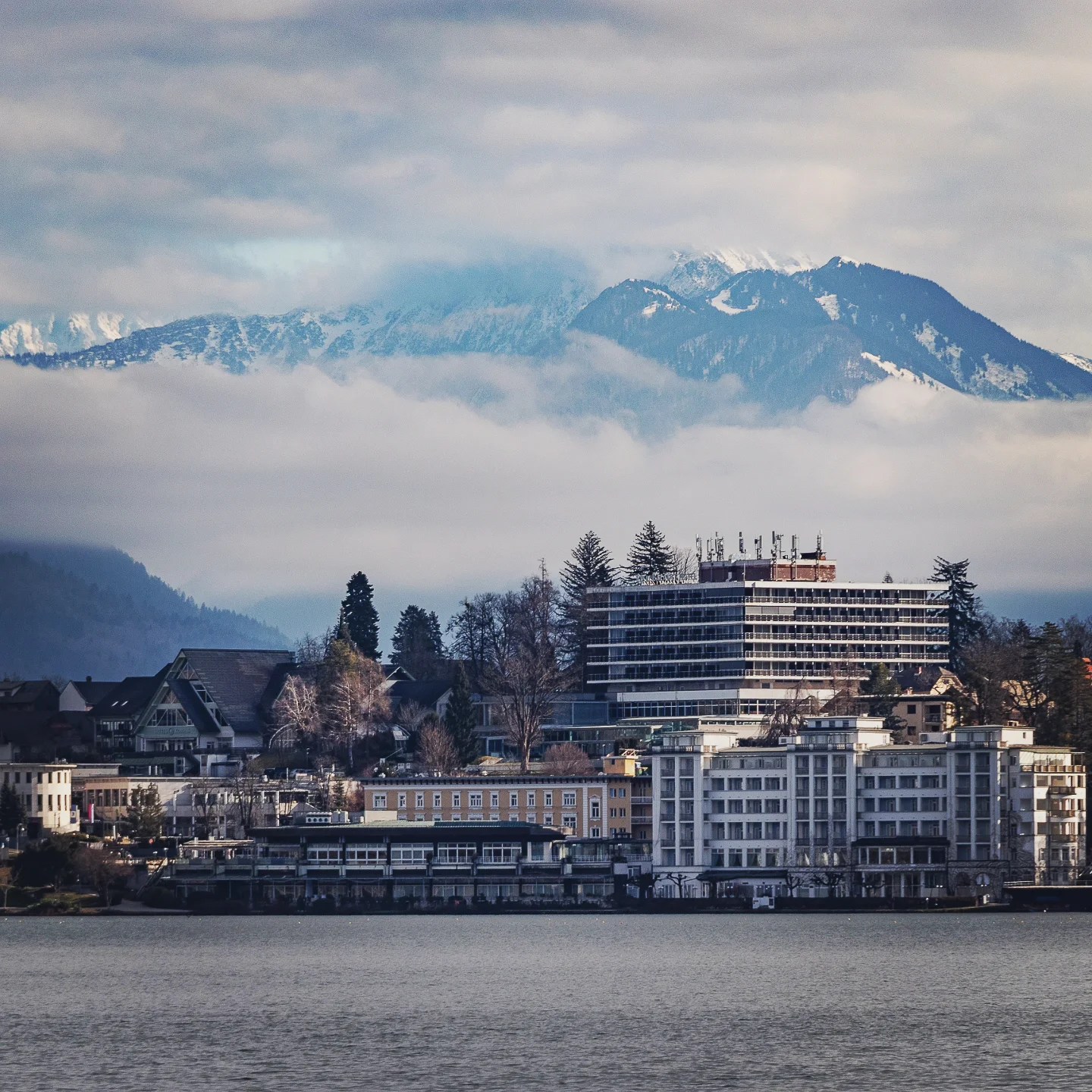 Cluster of lakeside hotels and buildings in Bled town with snowy mountains rising behind and low clouds drifting across the peaks.