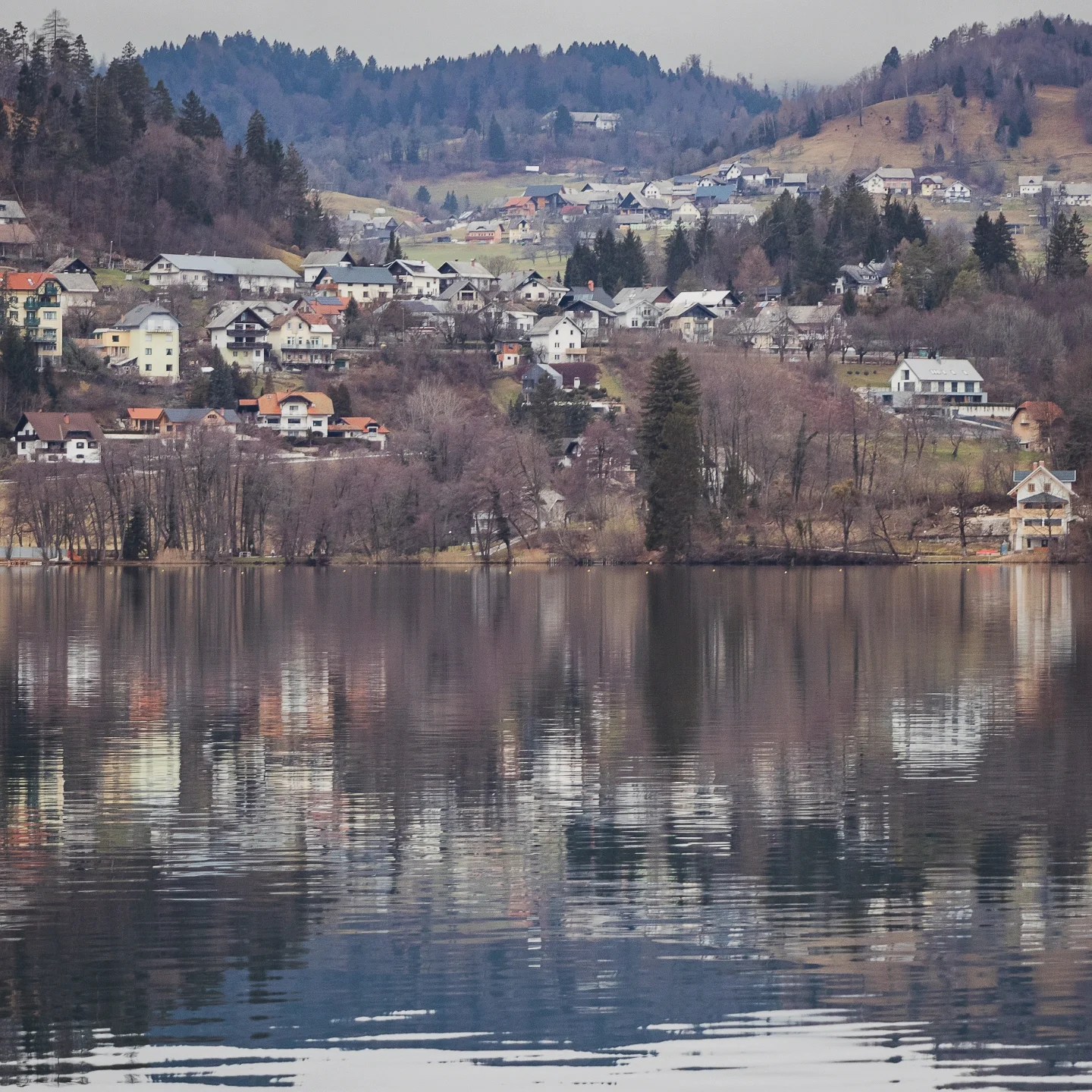 Quaint village homes dotting the hilly landscape above Lake Bled, their reflections mirrored in the still lake water below.
