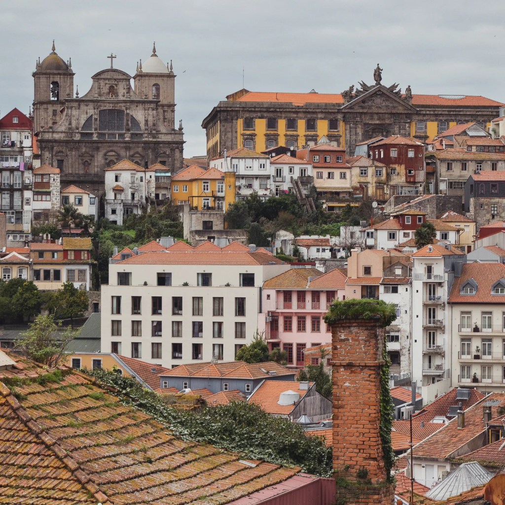 Rooftop view with moss-covered chimneys and a distant view of Porto Cathedral and other historic buildings.