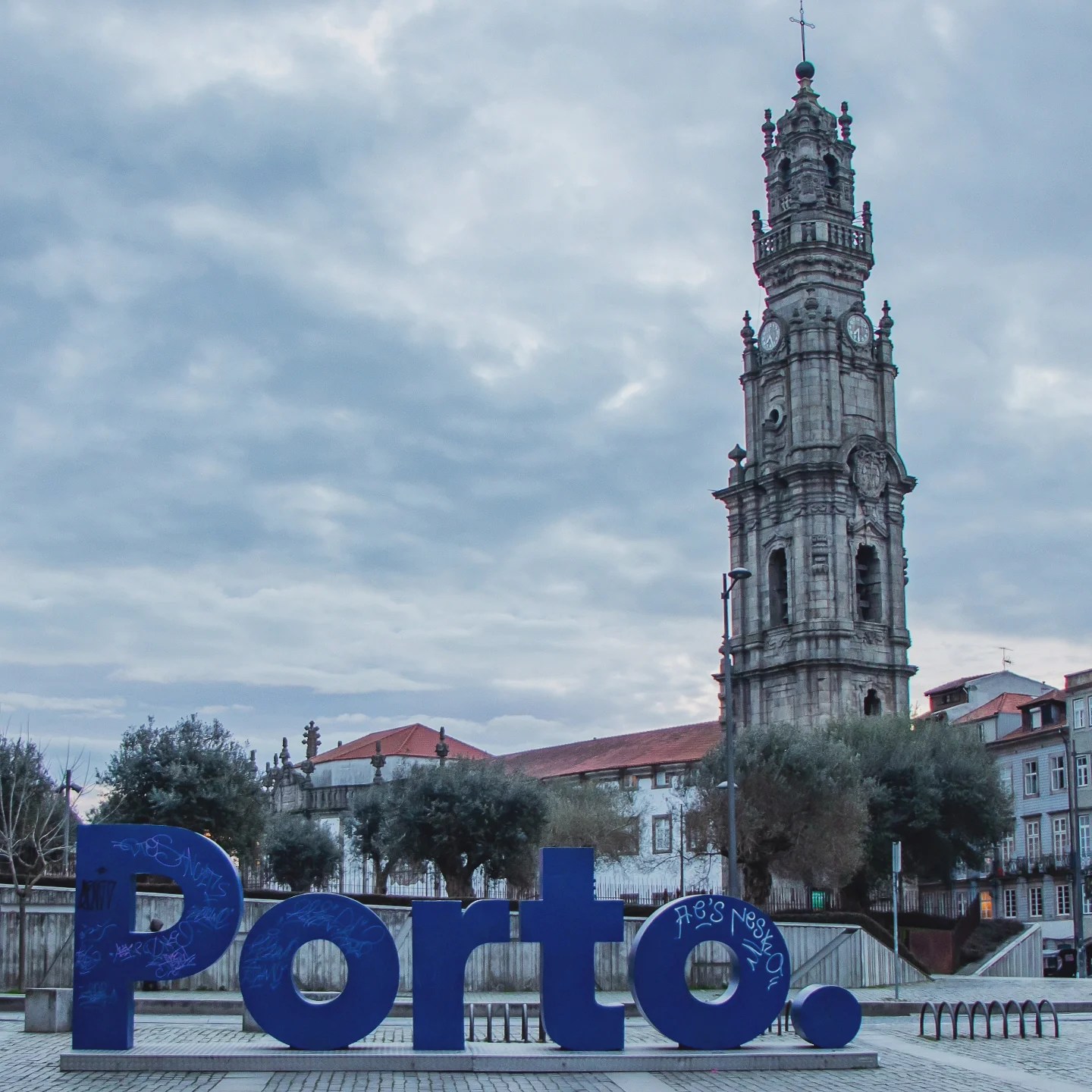 Porto’s iconic Clérigos Tower with a large blue Porto sign in the foreground under a cloudy sky.
