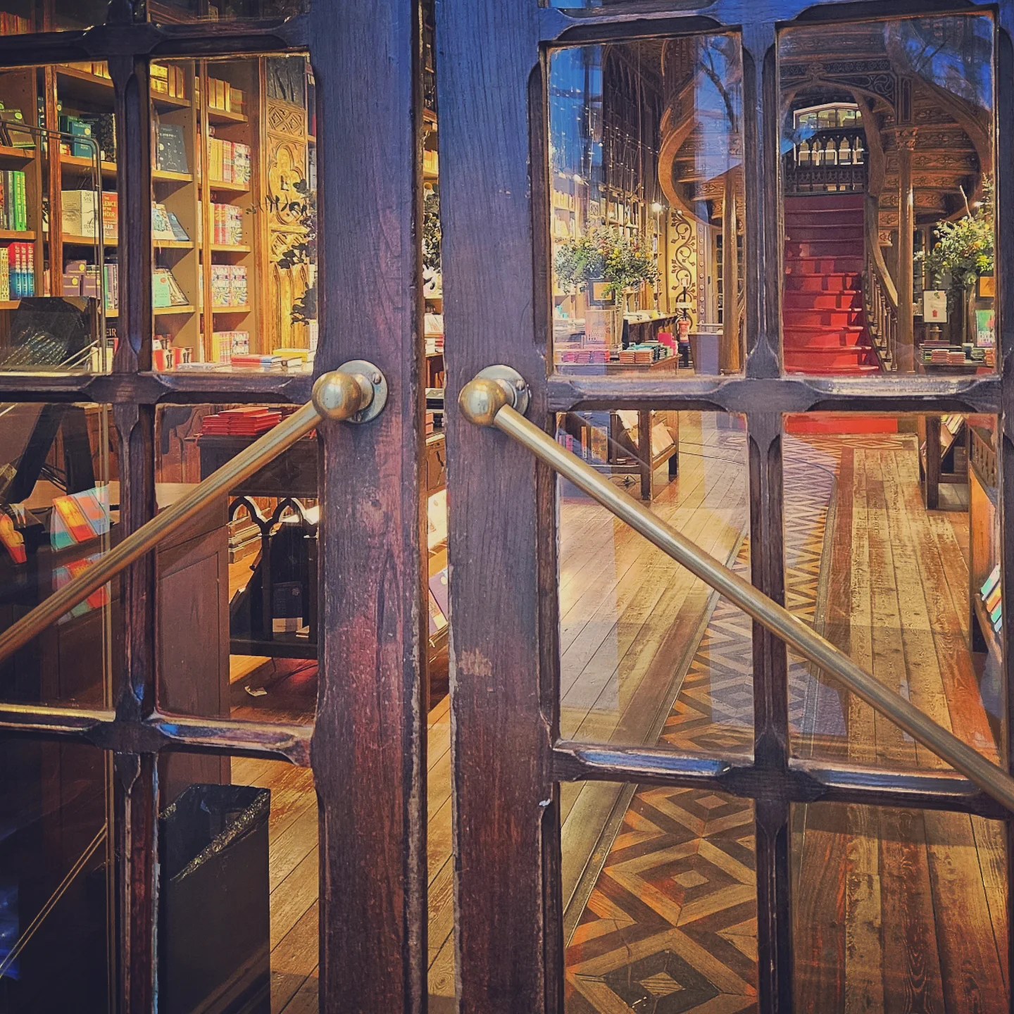 Wooden double doors with glass panes and brass handles, offering a view into the ornate interior of Livraria Lello bookstore in Porto, Portugal, with its famous red staircase.