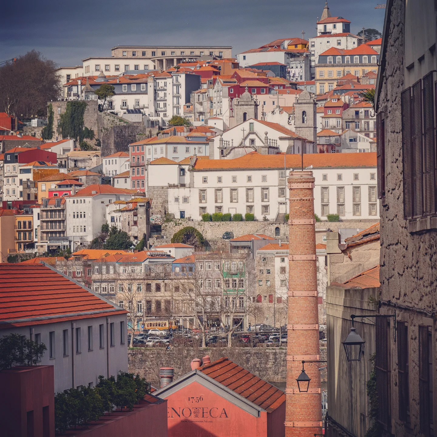 A layered cityscape of Porto with colorful houses, historic buildings, and a tall brick chimney stack from the wine cellars.