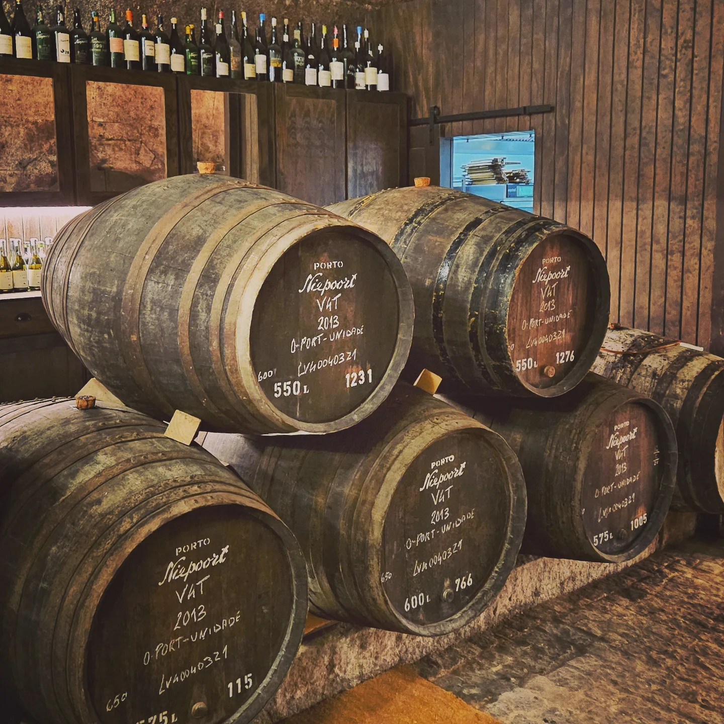 Stacked wooden wine barrels labeled Niepoort 2013 Vintage Port, with bottles lining the shelves behind them.
