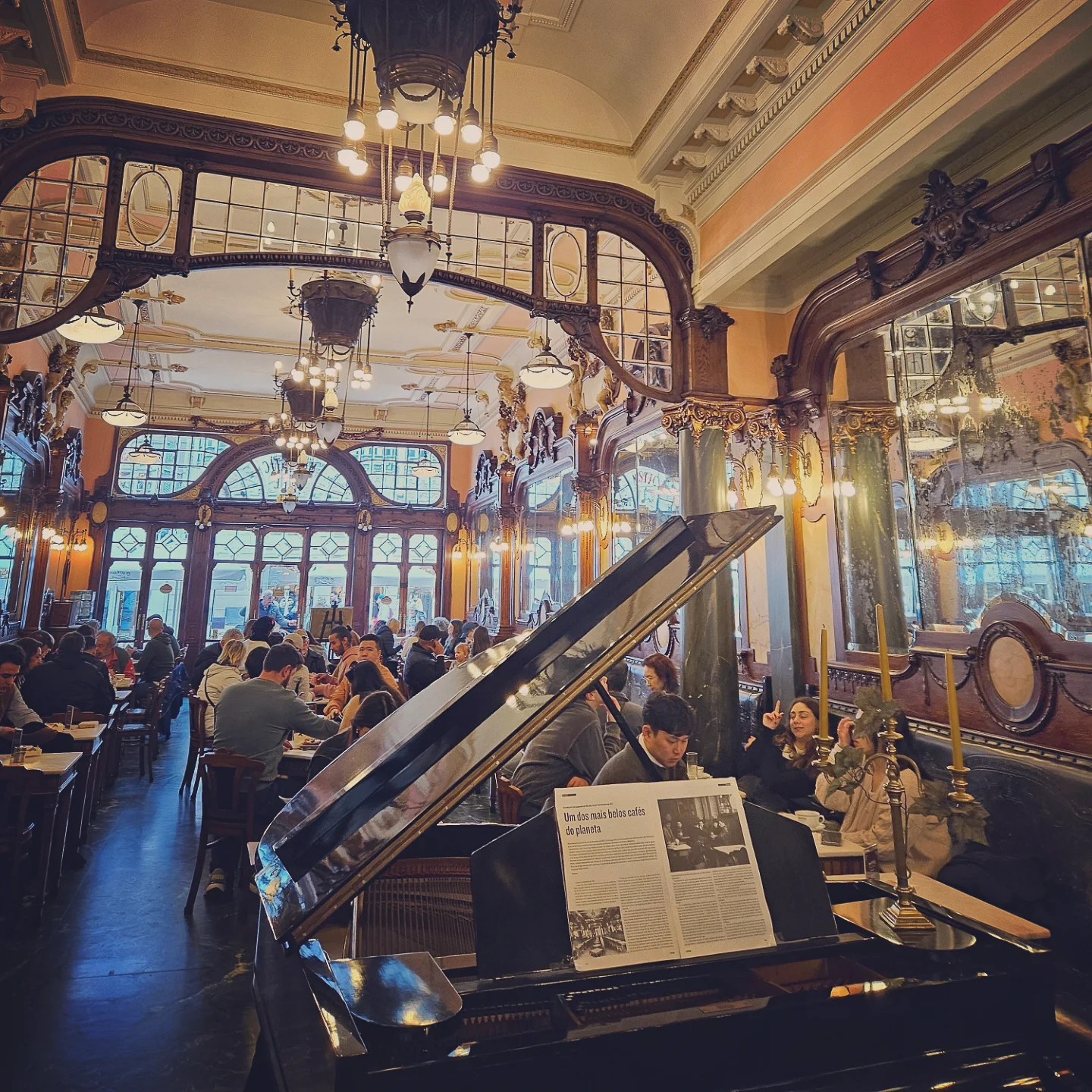 Majestic Café’s grand dining room with a central black piano, Art Nouveau décor, and guests seated at wooden tables.