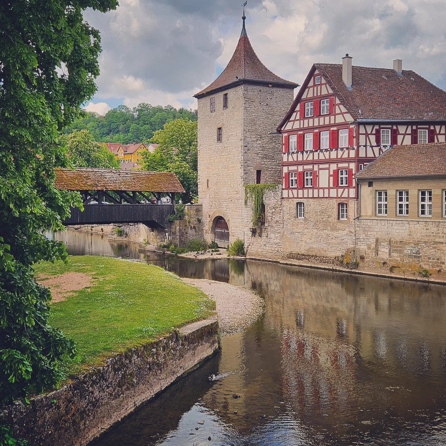 A scenic view of the Sulfersteg, a moss-covered wooden bridge crossing the Kocher River in Schwäbisch Hall, Germany. Beside it stands a historic stone tower and a traditional half-timbered house, framed by lush greenery and dramatic spring clouds.
