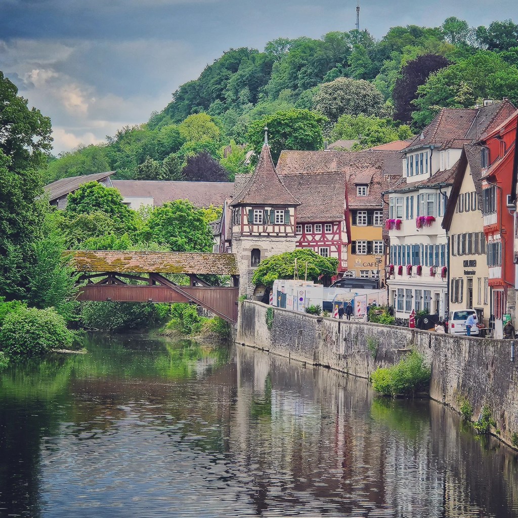 A scenic riverside view in Schwäbisch Hall, Germany, featuring colorful half-timbered buildings, a quaint café, and a covered wooden bridge crossing the Kocher River. Lush greenery and a hillside forest create a peaceful backdrop, while the river reflects the charming architecture.