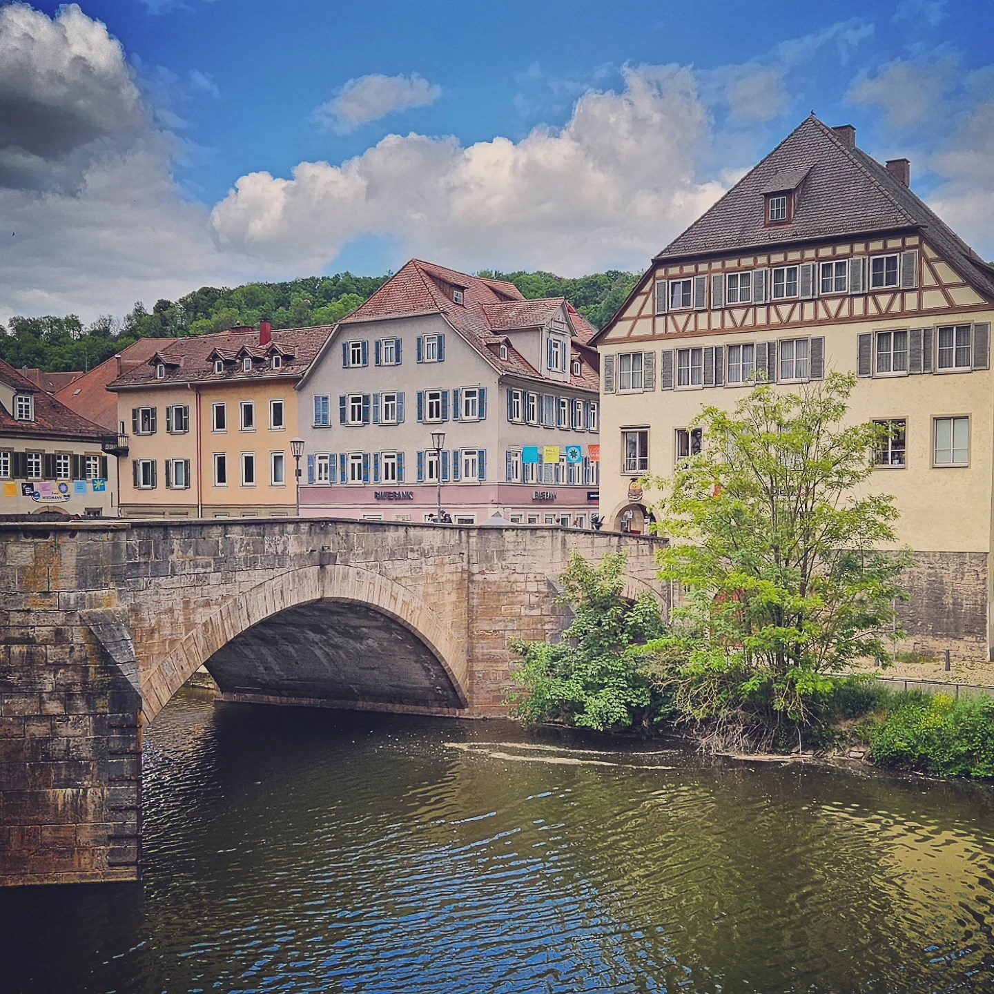 Stone arch bridge crossing the Kocher River in Schwäbisch Hall, with pastel-colored historic buildings and timber-framed architecture in the background under a bright blue sky with fluffy clouds.