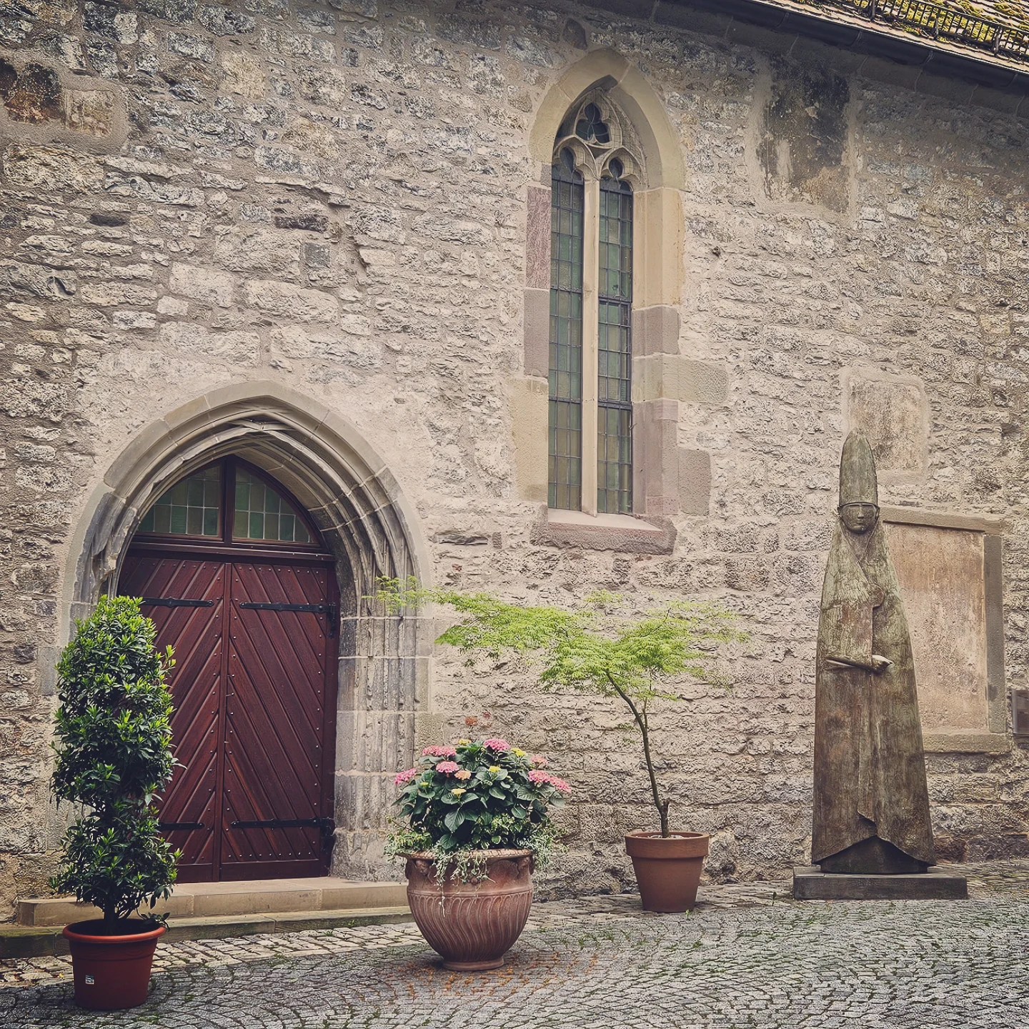 Stone courtyard of the Johanniterkirche in Schwäbisch Hall with arched wooden door, gothic-style windows, potted plants, and a tall bronze statue of a bishop-like figure.