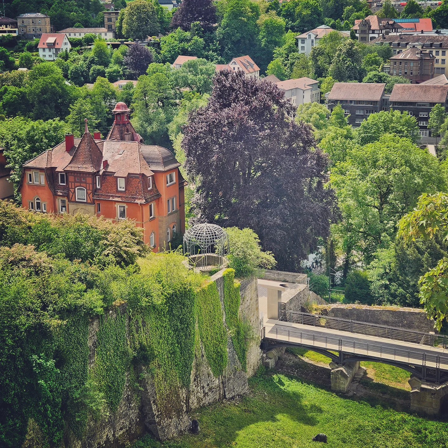 A vibrant view of a red-roofed historic house perched above an ivy-covered stone wall in Schwäbisch Hall, Germany. A small metal gazebo and a modern footbridge connect the structure to a lush green park below, with leafy trees and rooftops of the surrounding town visible in the background.