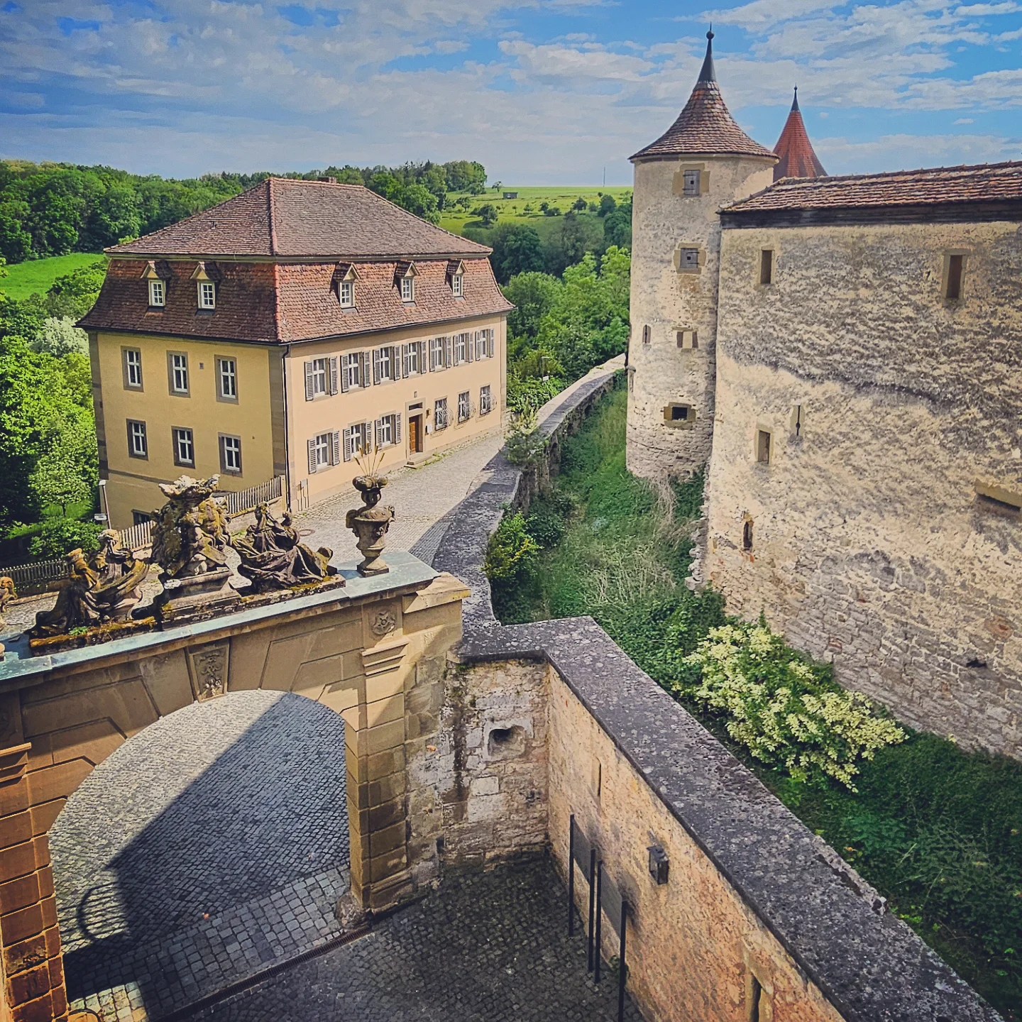 A scenic view of Großcomburg Monastery near Schwäbisch Hall, Germany, featuring a fortified stone wall with round towers, an ornate entrance gate topped with statues, and a yellow Baroque-style building overlooking the lush green countryside under a blue sky.