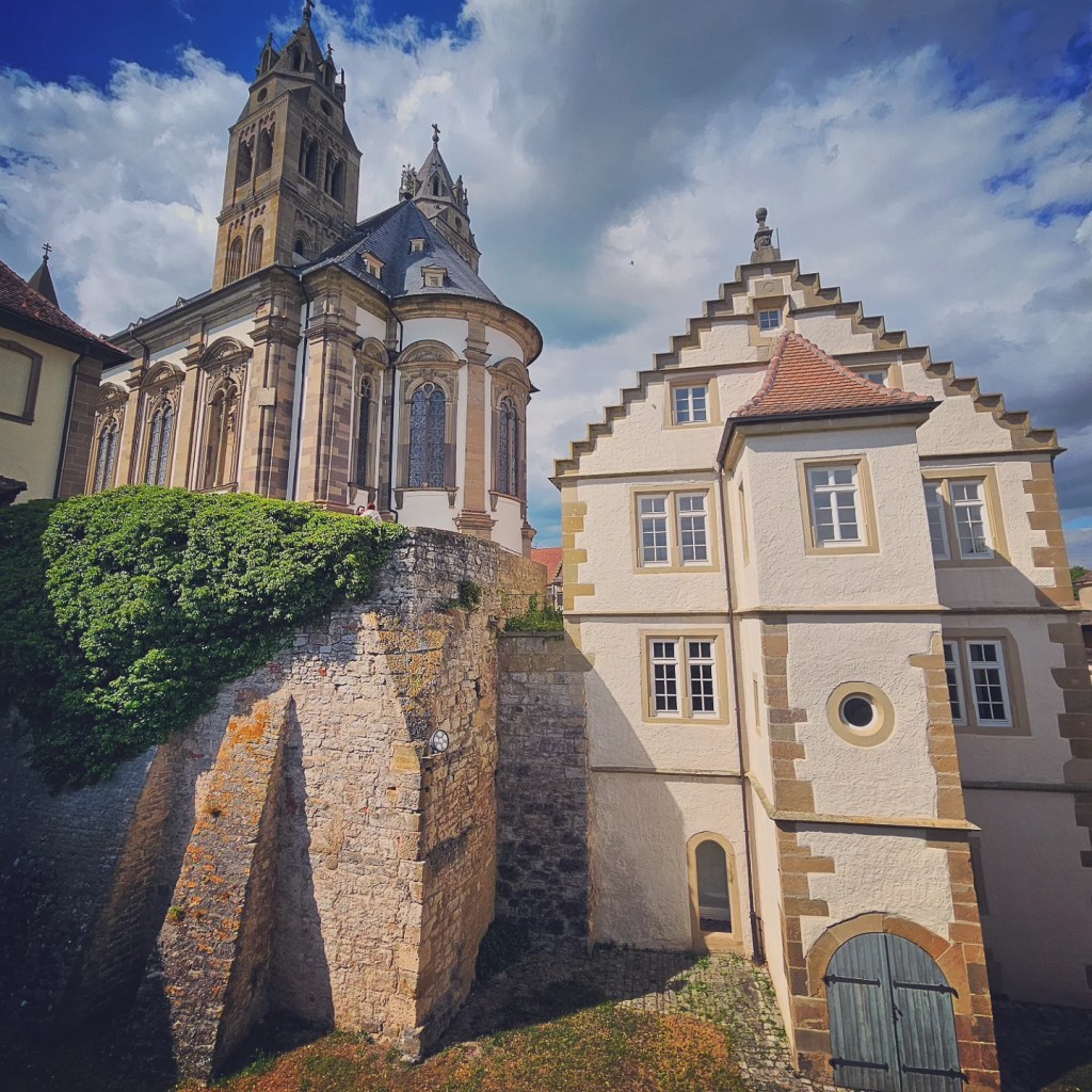 View of the historic Großcomburg Monastery with its intricate architecture and surrounding greenery with clouds and a blue sky in the background.