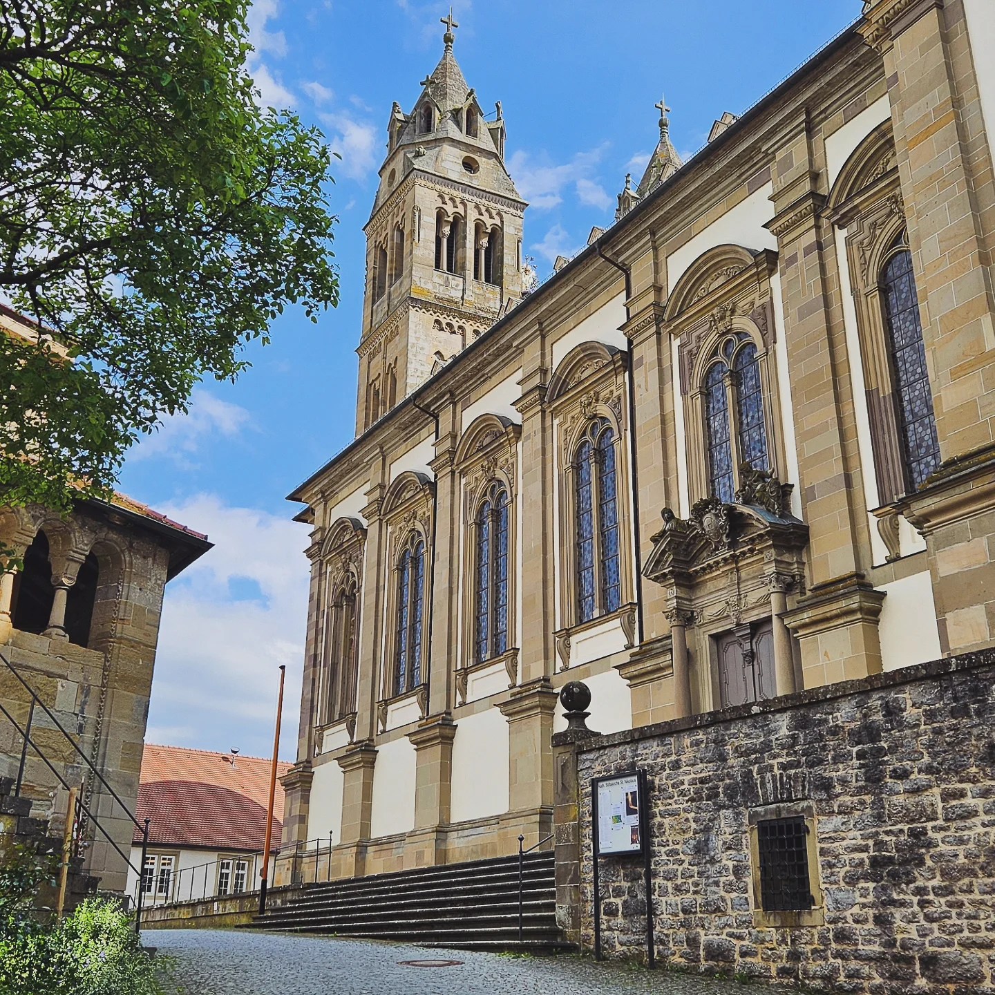 A striking view of the Collegiate Church of St. Nicholas at Großcomburg Monastery, featuring a tall Romanesque bell tower and a grand facade with arched stained-glass windows and ornate stonework. The church is set against a bright blue sky with sunlight casting shadows across the stone steps and courtyard below.