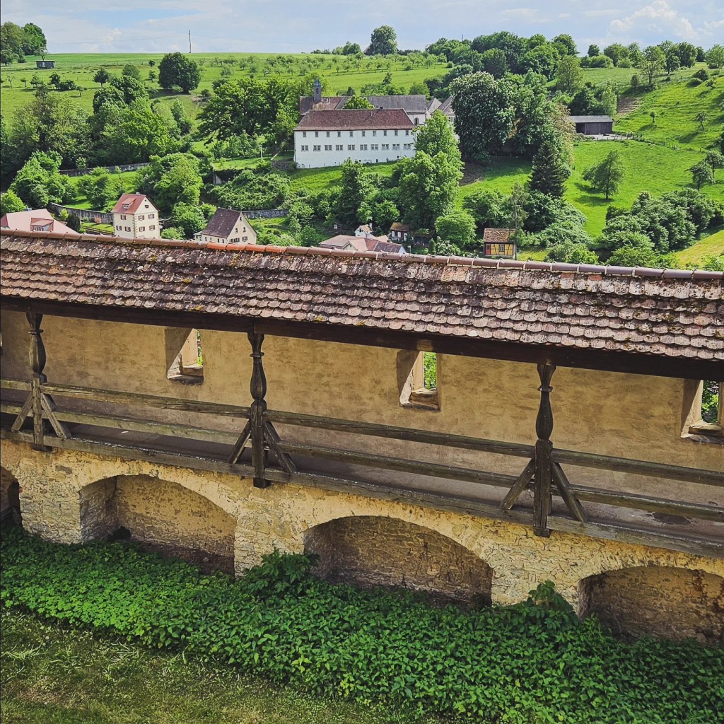 View of the fortified wall of Großcomburg Monastery with lush green hills and a village in the background.