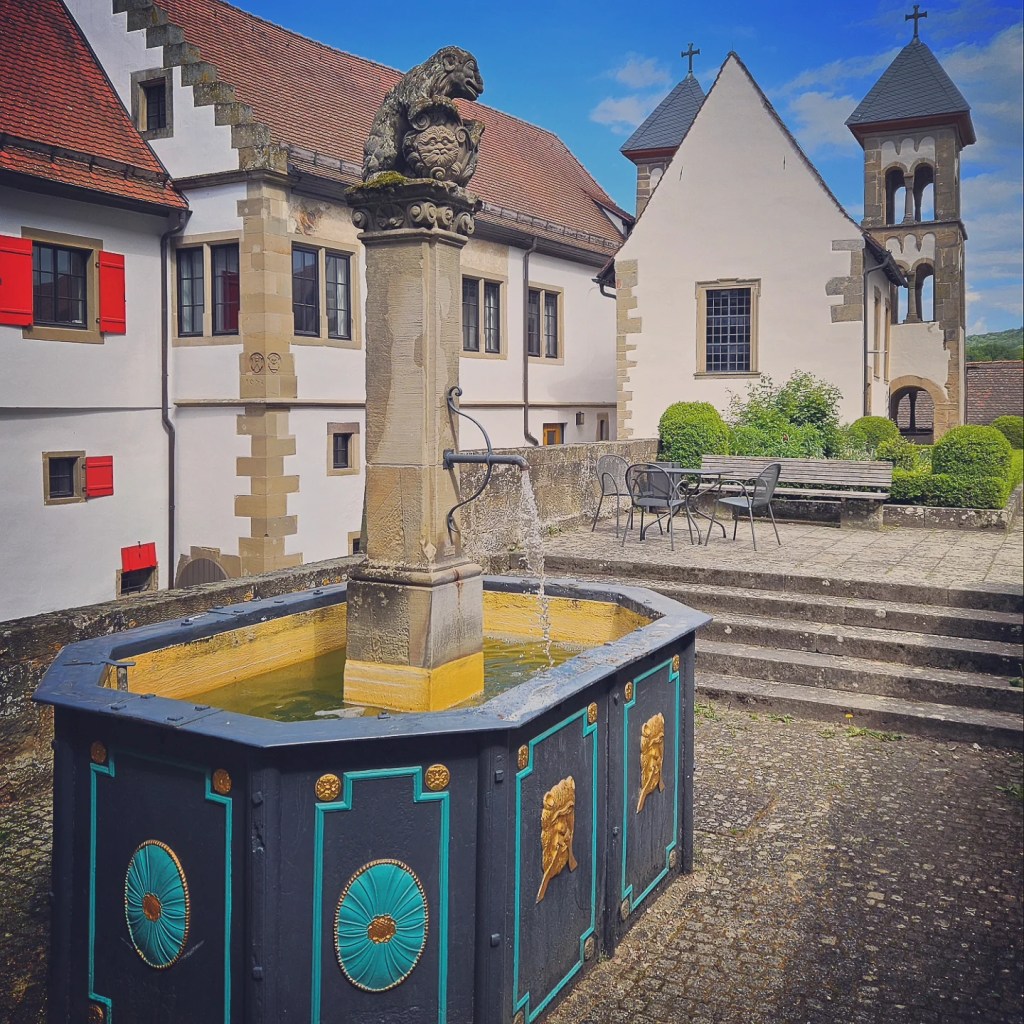A historic courtyard featuring a decorative fountain with a stone sculpture at the top. Surrounding buildings display half-timbered architecture and vibrant red shutters against a clear blue sky.