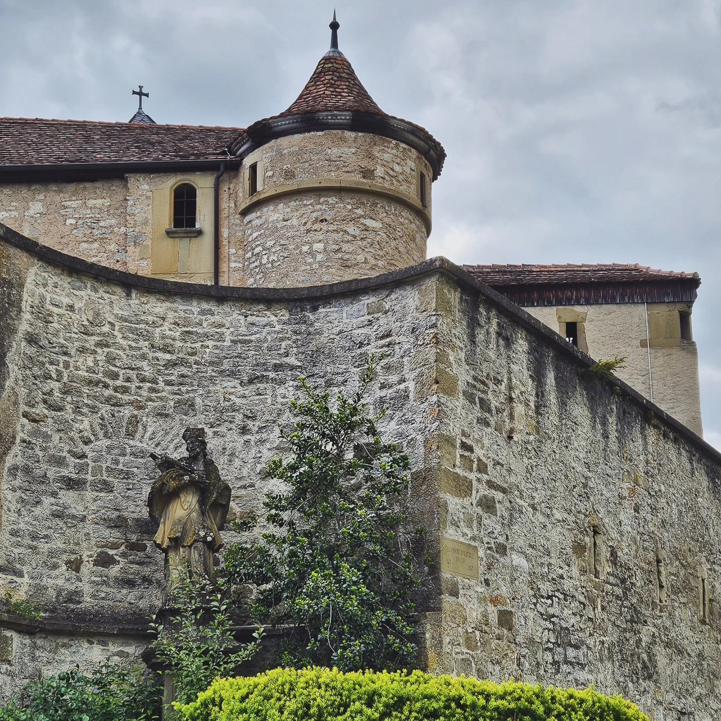 A dramatic stone wall curves around a statue of a robed figure at Großcomburg Monastery in Germany. Above the wall, a round turret with a conical red-tiled roof rises from the fortress-like structure, set against a cloudy sky. Shrubs and greenery grow around the base, softening the medieval architecture.