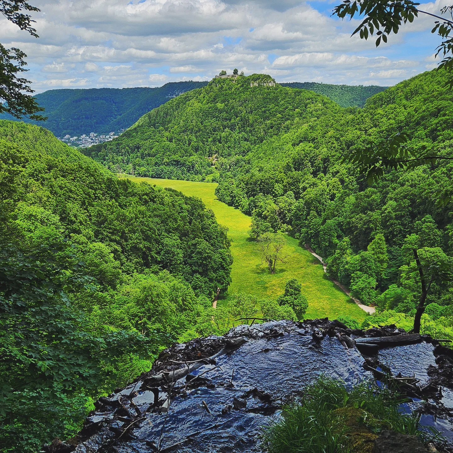 View from the top of Urach Waterfall, looking out over the lush green Maisental valley with forested hills and castle ruins in the distance beneath a partly cloudy sky.