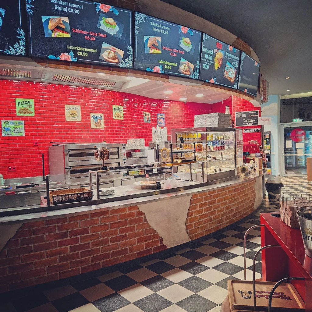 A diner-style counter with a red tile wall, food warmers, menus, and pastries on display.
