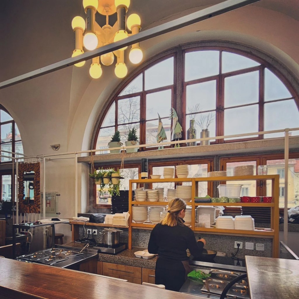 A woman preparing food behind a counter at a warm, modern restaurant with arched windows and stacked white plates.