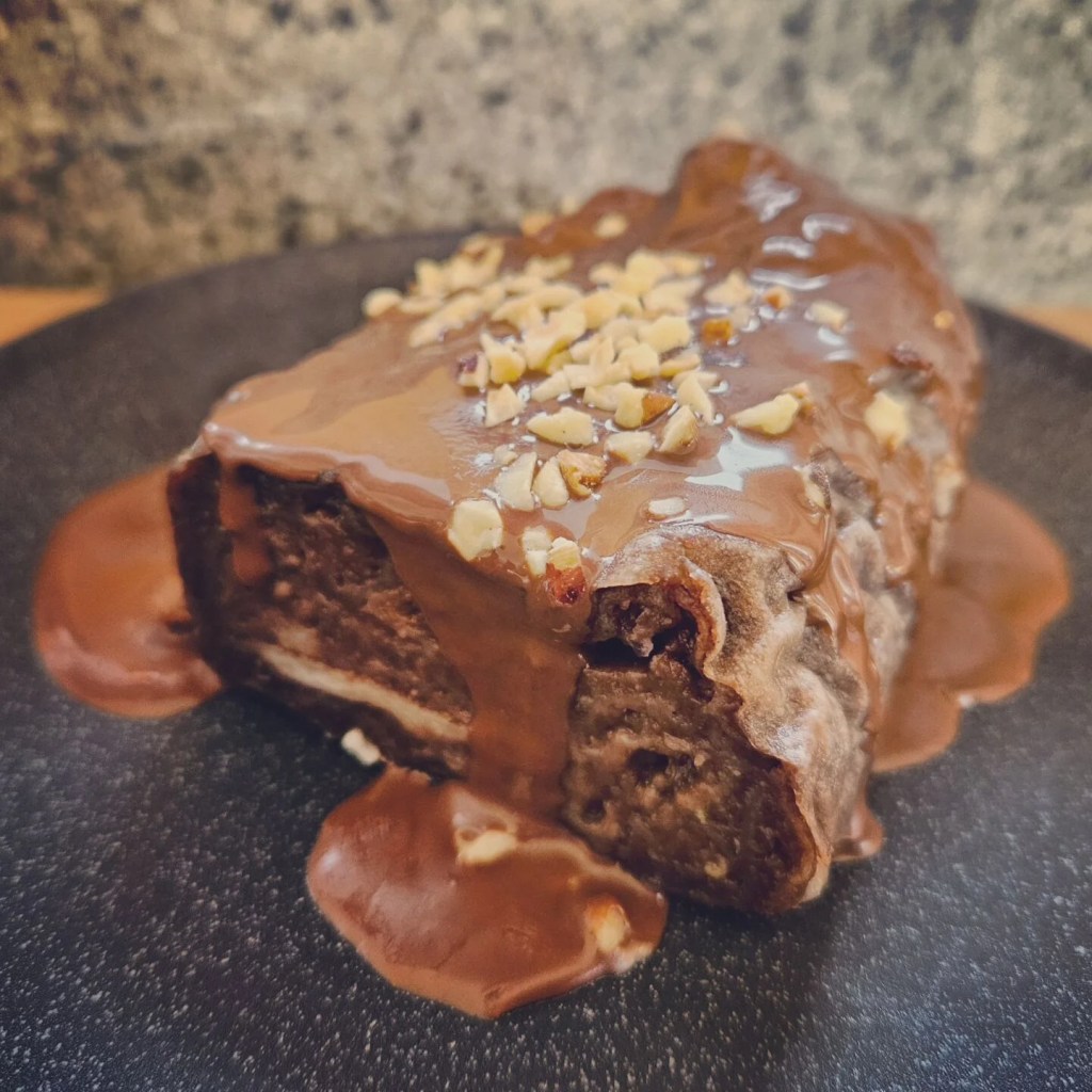 A rich slice of chocolate dessert topped with chopped nuts and drizzled with sauce on a black speckled plate.