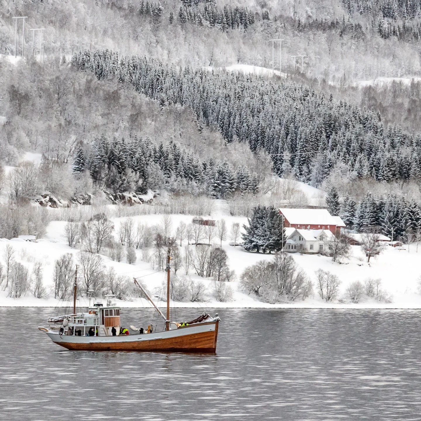 Classic wooden boat cruising past snow-covered hills and forested shoreline.