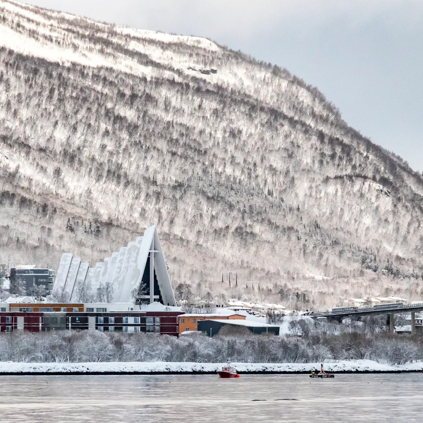 A striking view of the Arctic Cathedral with its modern, angular design nestled beneath a snow-covered mountain, with colorful buildings and fishing boats in the foreground.