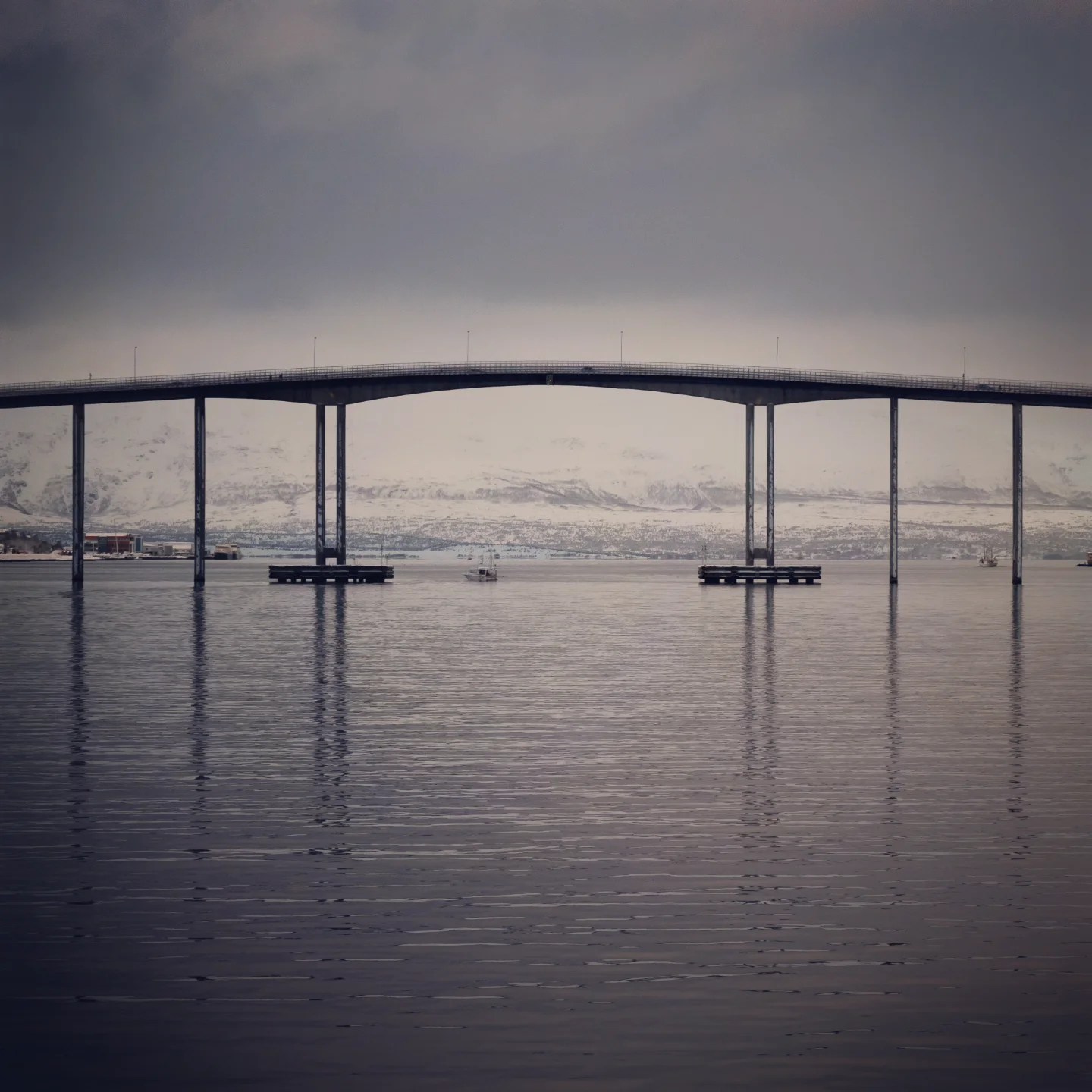 A minimalist, moody image of Tromsø Bridge stretching across a still, icy fjord under a cloudy winter sky.