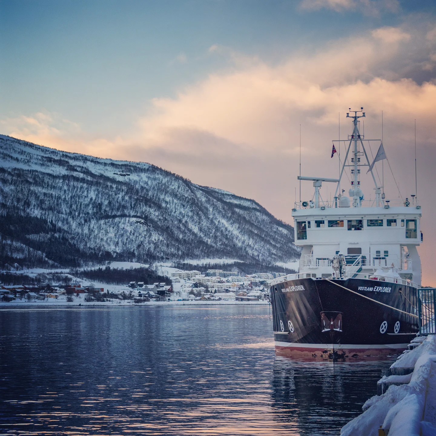 The Vestland Explorer moored along the Tromsø waterfront with a scenic snowy mountain and soft, colorful winter sky in the background.
