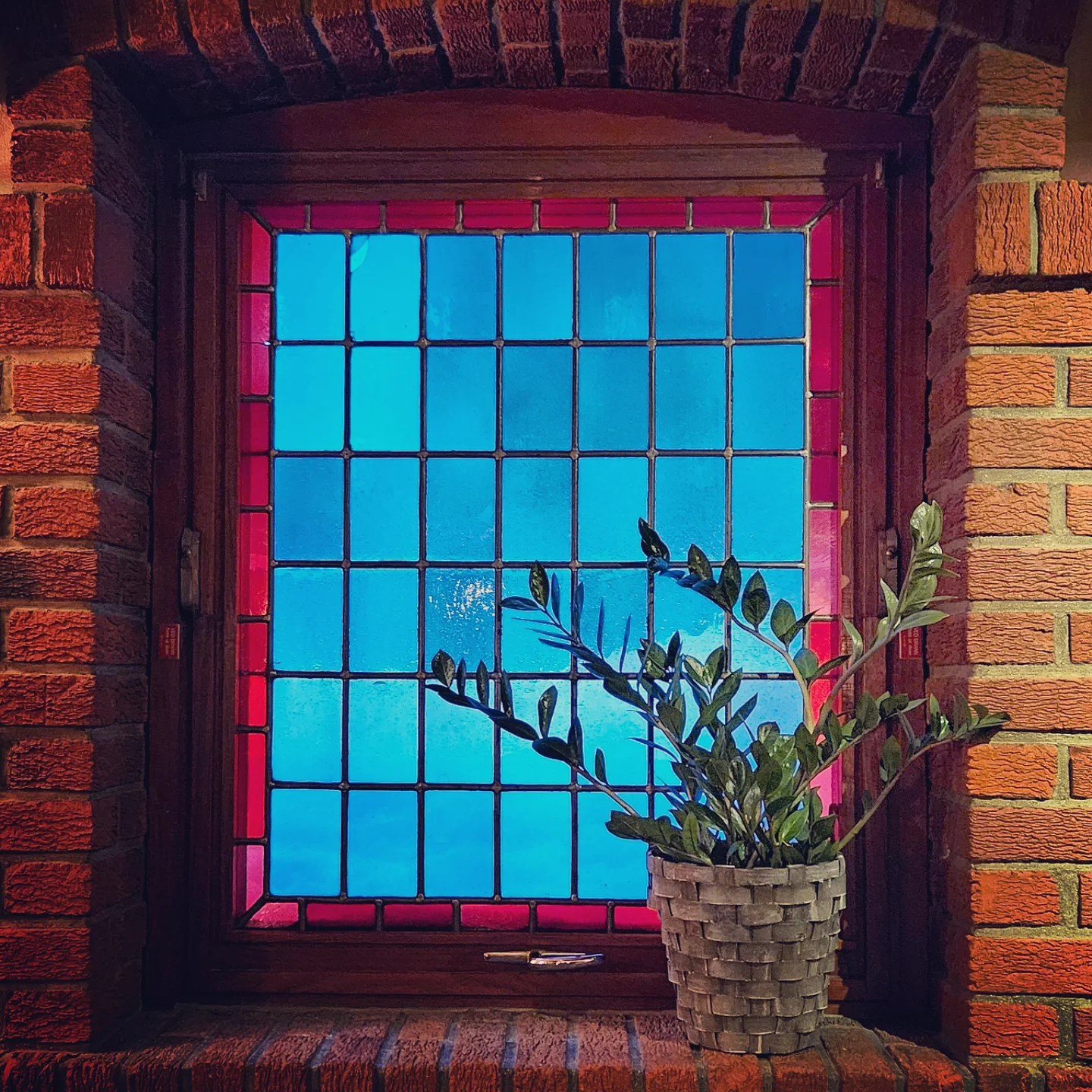 A vibrant stained glass window framed by red brick, with a leafy potted plant on the windowsill.