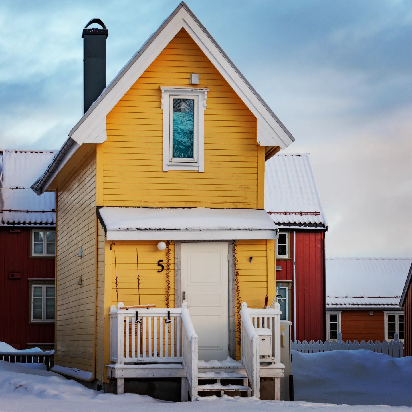 A charming narrow yellow house with white trim and a snowy roof, nestled among colorful wooden homes under a soft winter sky.