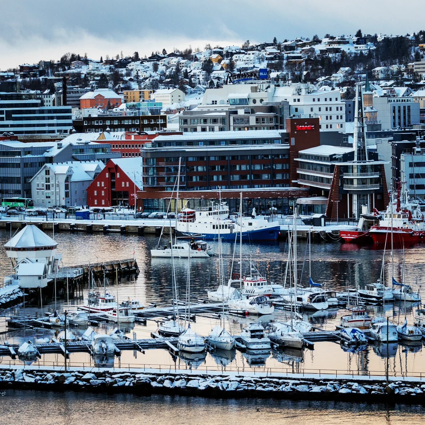 View of Tromsø's modern harbor with boats docked in the marina and snow-covered buildings clustered against a hillside backdrop.