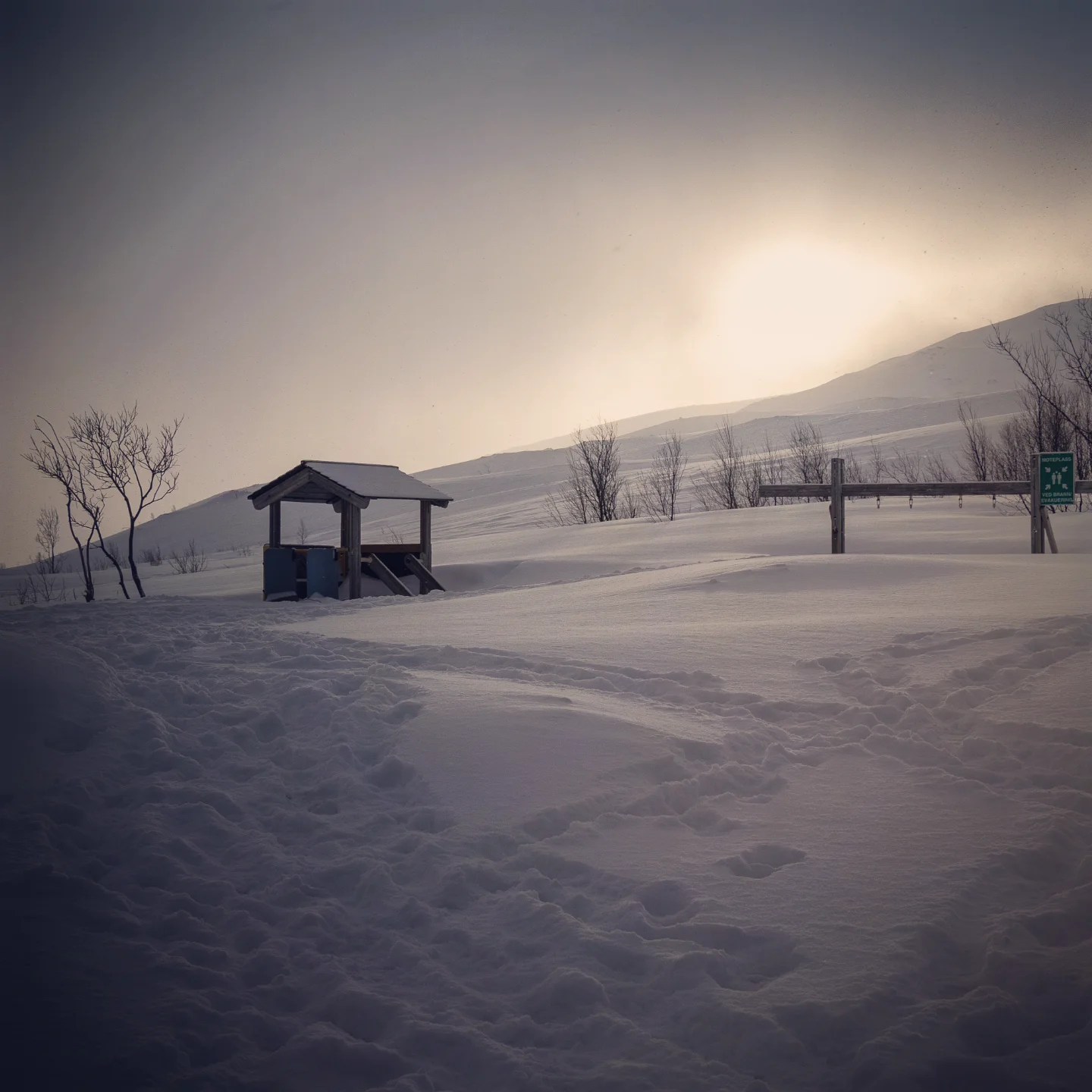 Desolate wooden shelter surrounded by deep snow in a wintry landscape, with pale sunlight breaking through overcast skies.
