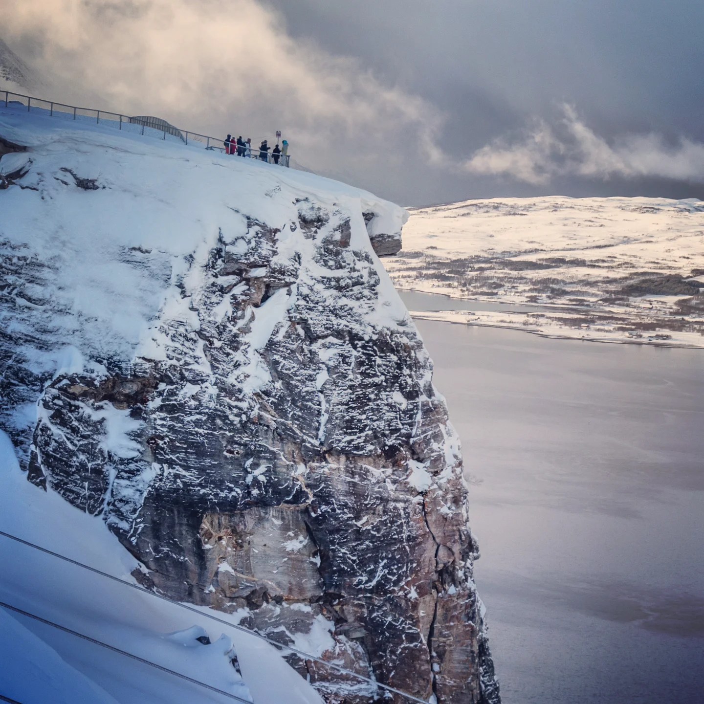 Snow-covered cliff edge with people gathered at a lookout point, peering over a dramatic fjord under heavy clouds.