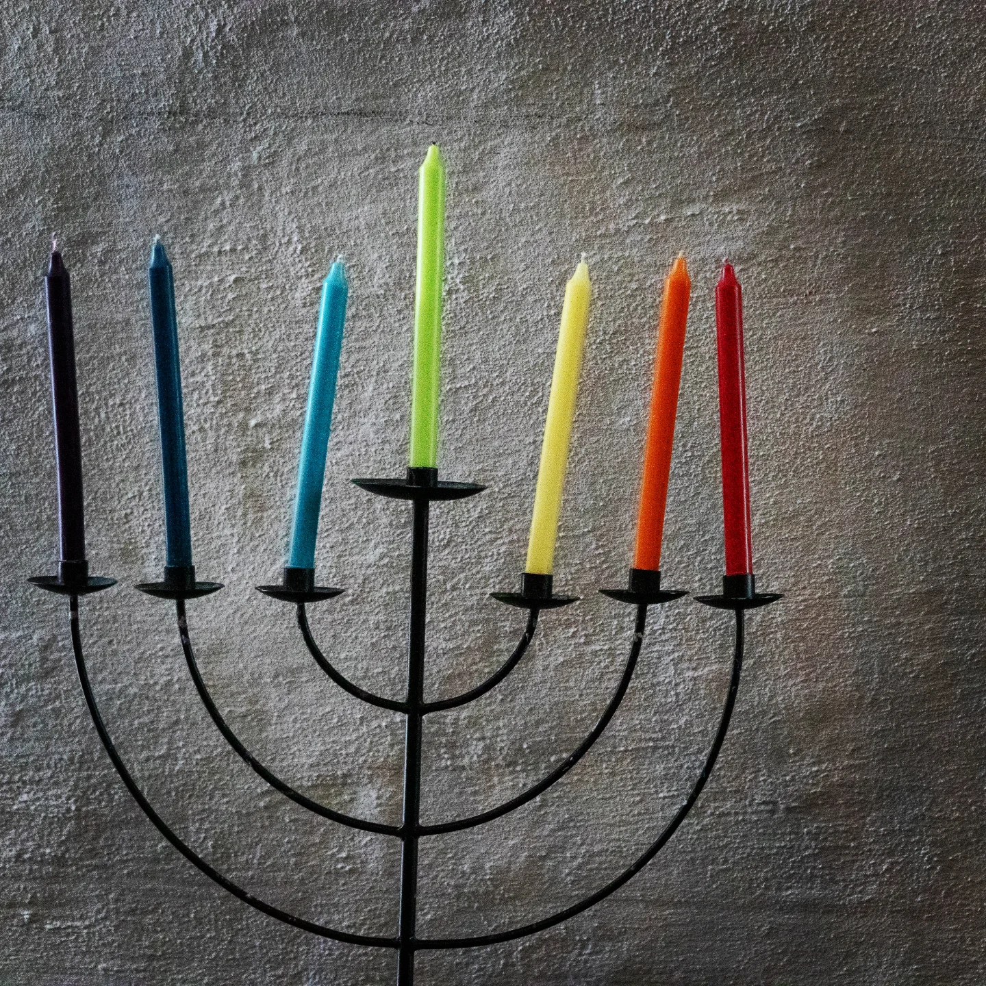 Rainbow-colored candles in a black metal menorah, set against a textured gray wall.