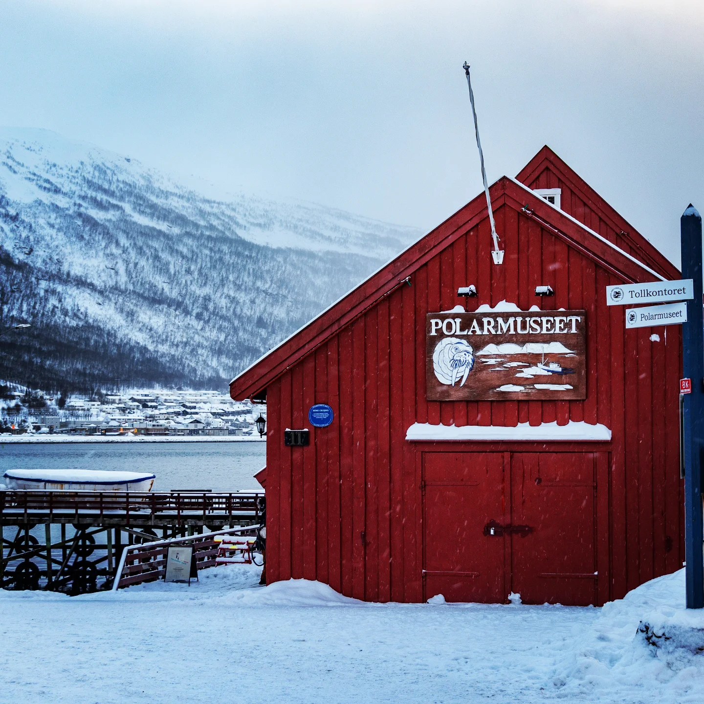The iconic red wooden facade of the Polarmuseet in Tromsø, blanketed in snow with fjord and mountains in the background.