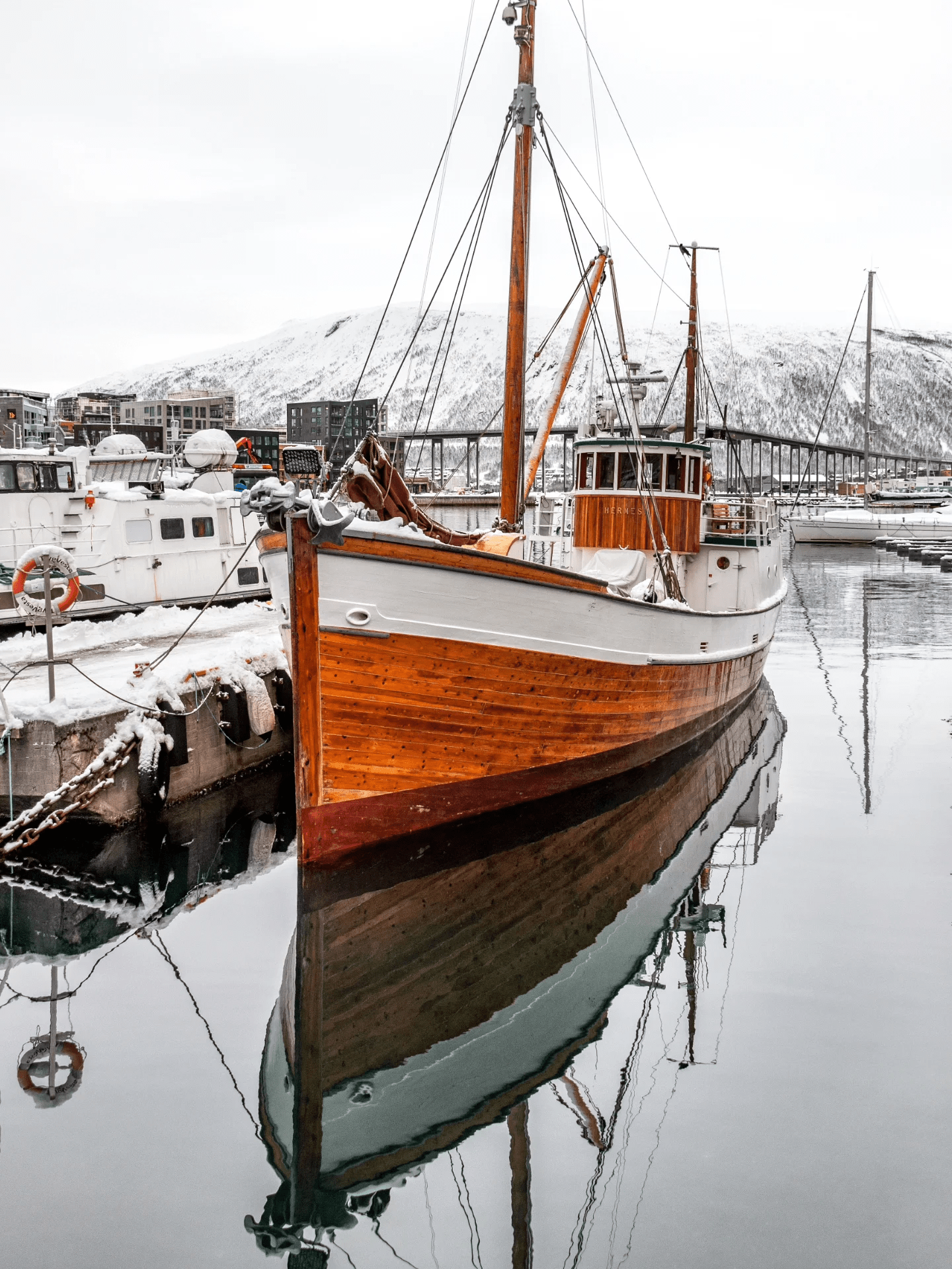 A beautifully preserved wooden fishing vessel docked in a snow-covered harbor, reflecting perfectly in the still water, with snow-capped mountains in the background.