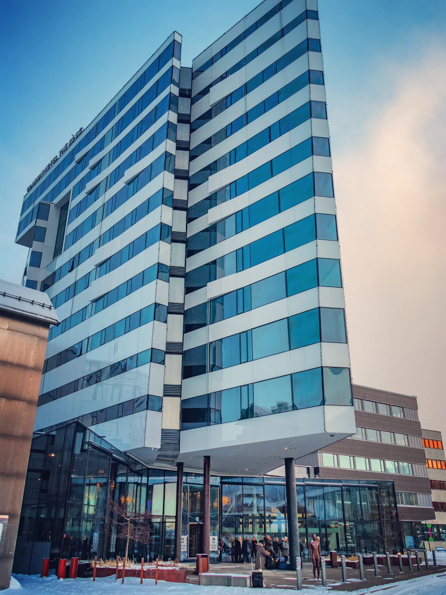 A modern glass-fronted hotel in Tromsø rising into a blue winter sky, its entrance busy with warmly dressed guests arriving and departing.