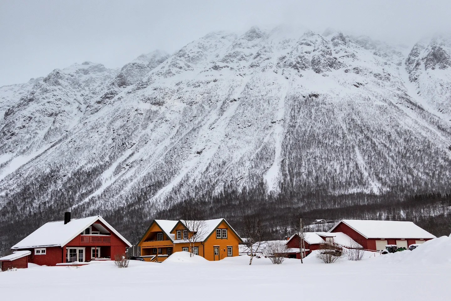 Brightly painted wooden houses in red and yellow sit surrounded by untouched snow, dwarfed by towering white mountains rising steeply behind them.