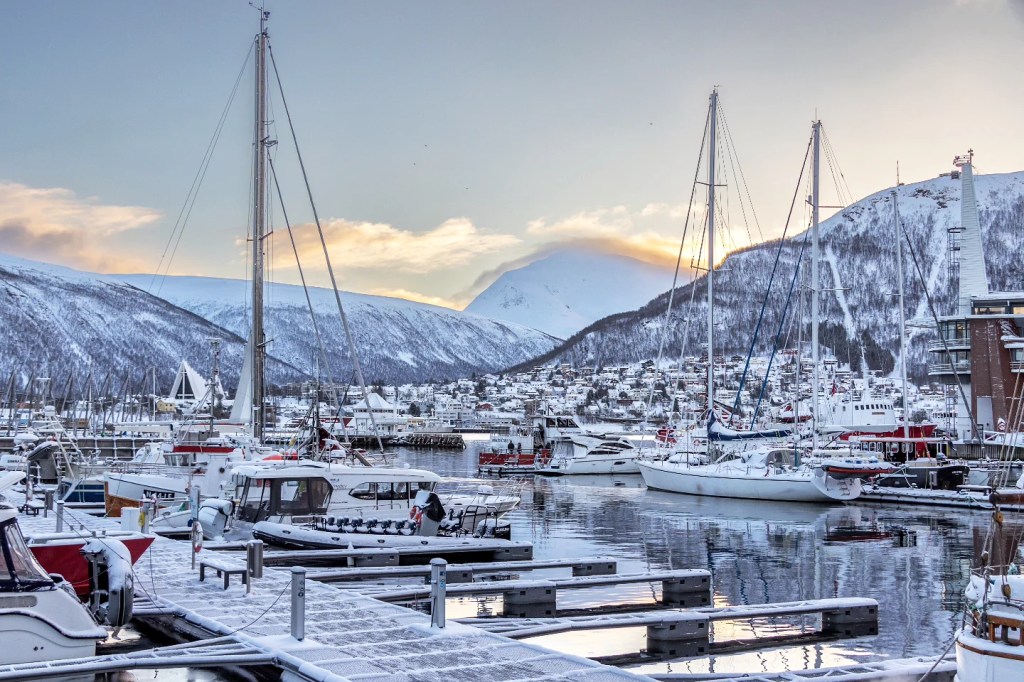 Boats and yachts docked in Tromsø’s marina as the first golden rays of sunrise peek over snow-laced peaks, casting a warm glow on the icy water.