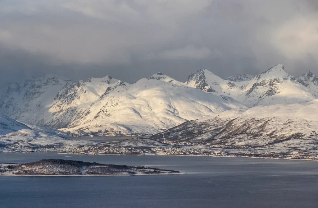 A dramatic Arctic landscape of rugged, snow-covered mountains and dark fjords bathed in golden afternoon light under a brooding sky.