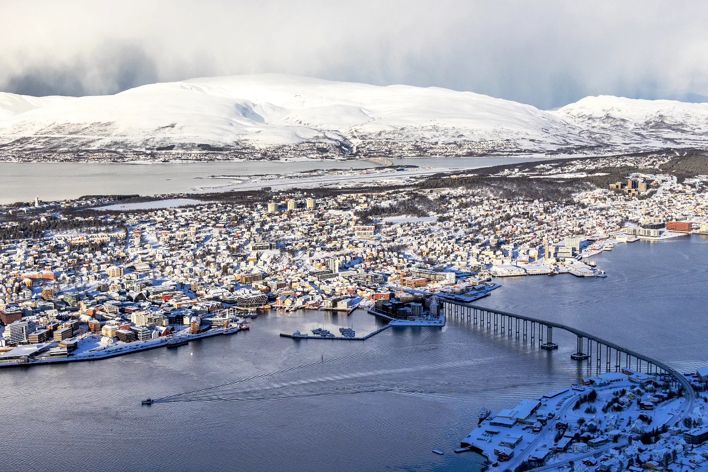 A sweeping aerial view of Tromsø, highlighting the elegant curve of the Tromsø Bridge, colorful buildings, and surrounding fjords under a blanket of snow.