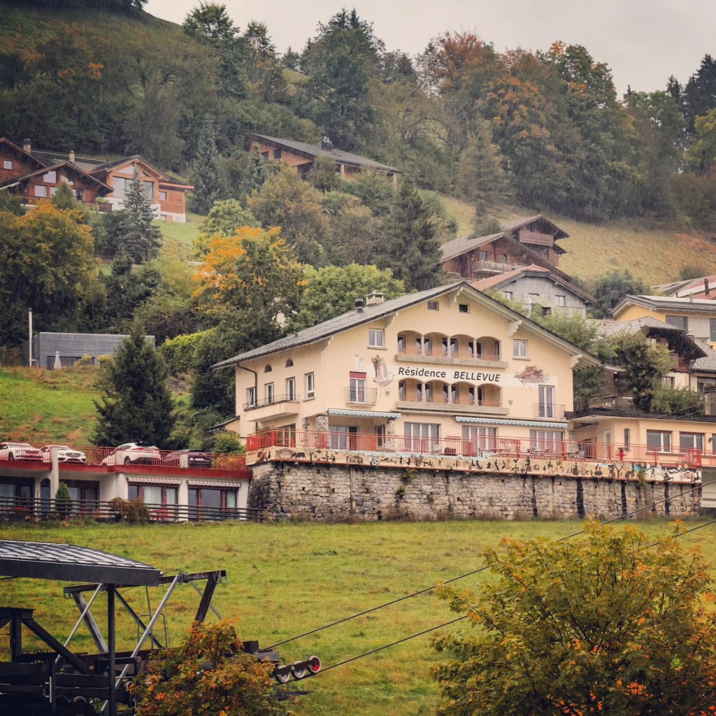 Yellow three-storey building marked “Résidence Bellevue” perched on a grassy hillside dotted with chalets.