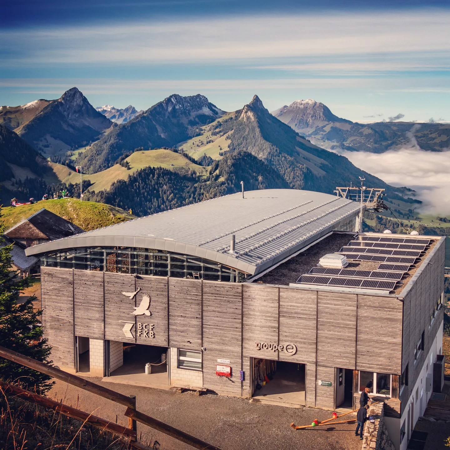 Modern timber-clad gondola terminal with solar panels; layers of rolling green mountains behind.